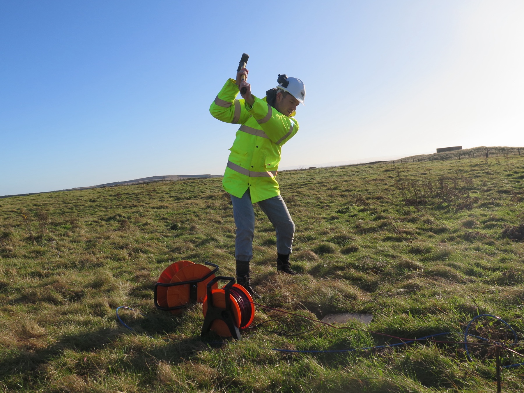 A surveyor carrying a geophysical instrument across open ground.